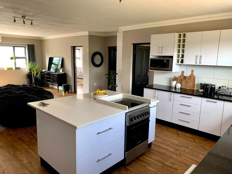 Modern open-plan kitchen featuring a central island with integrated hob and oven, white cabinetry, and contrasting black countertops, flowing into the living area.
