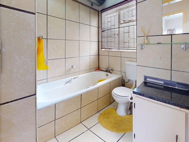 A dated bathroom featuring a built-in bathtub, toilet, and vanity with a dark countertop, finished with neutral wall and floor tiles.