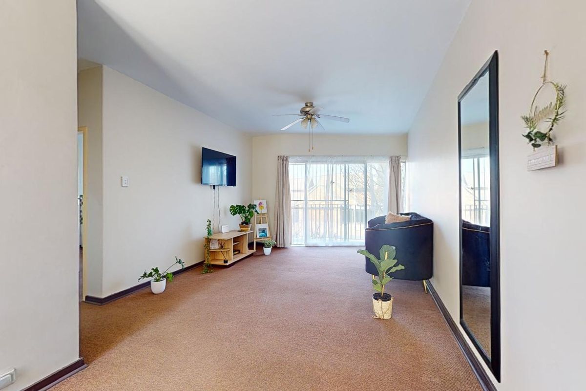 A spacious living room featuring neutral walls, carpeted flooring, a ceiling fan, and sliding glass doors leading to a balcony.
