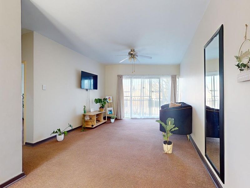 A spacious living room featuring neutral walls, carpeted flooring, a ceiling fan, and sliding glass doors leading to a balcony.