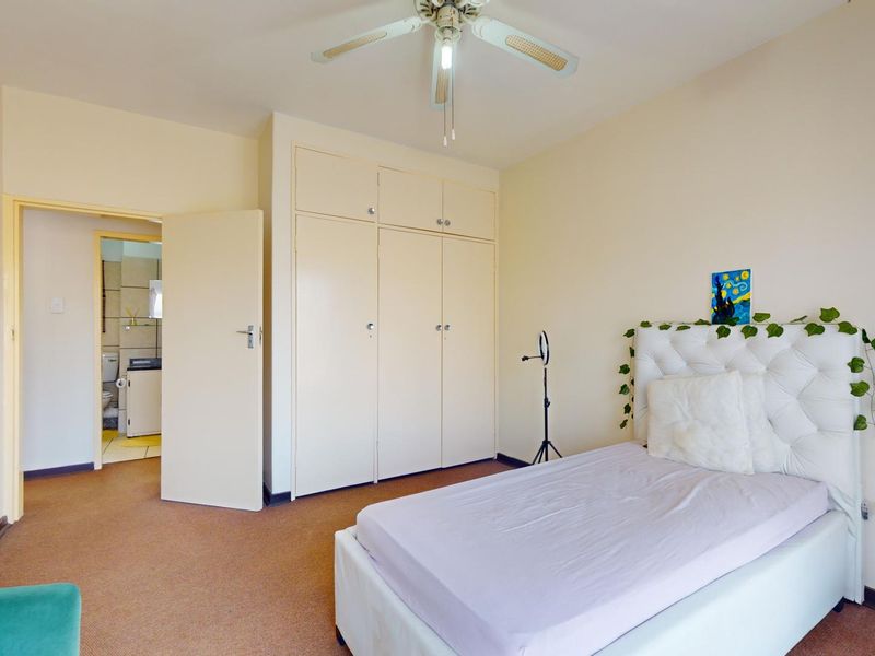 A bedroom featuring built-in cupboards, carpeted flooring, and a ceiling fan, with an adjoining bathroom visible through the doorway.