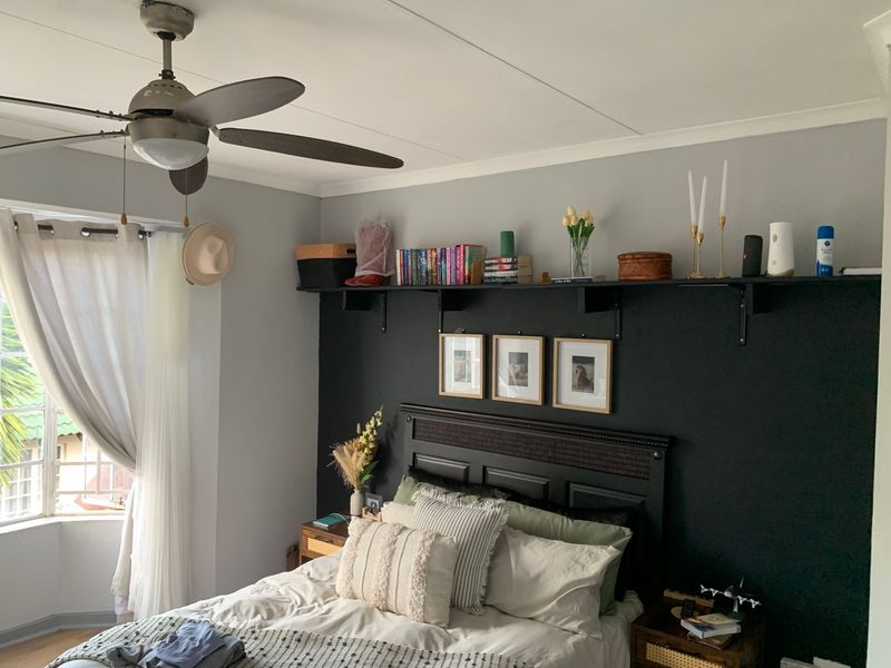 A well-maintained bedroom featuring a ceiling fan, a bay window with curtains, and a dark accent wall with built-in shelving.