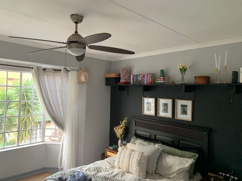 A well-lit bedroom featuring a ceiling fan, a large bay window, and a dark accent wall with integrated shelving.