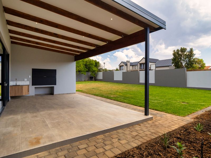 Covered patio featuring a built-in braai, exposed timber beams, and recessed lighting, overlooking a well-maintained garden and paved walkway.