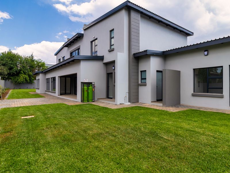 A well-maintained rear exterior view of a modern two-story house featuring a paved patio area, landscaped lawn, and contemporary grey facade finishes.