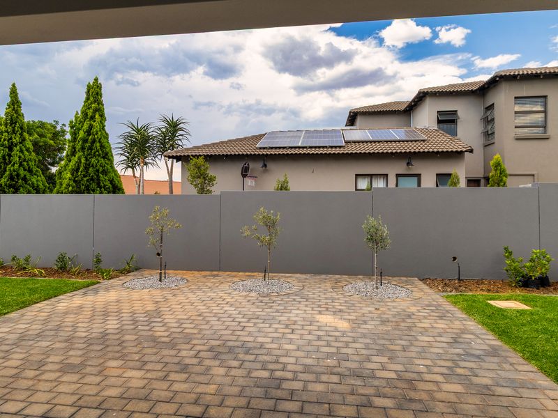 A well-maintained rear exterior view featuring a paved area, landscaped garden, boundary wall, and roof-mounted solar panels on the main dwelling.