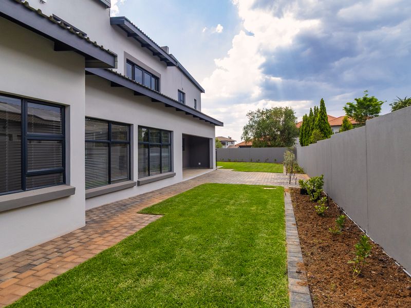 A well-maintained rear garden featuring a paved patio, manicured lawn, and a modern boundary wall, adjacent to the home's covered patio area.