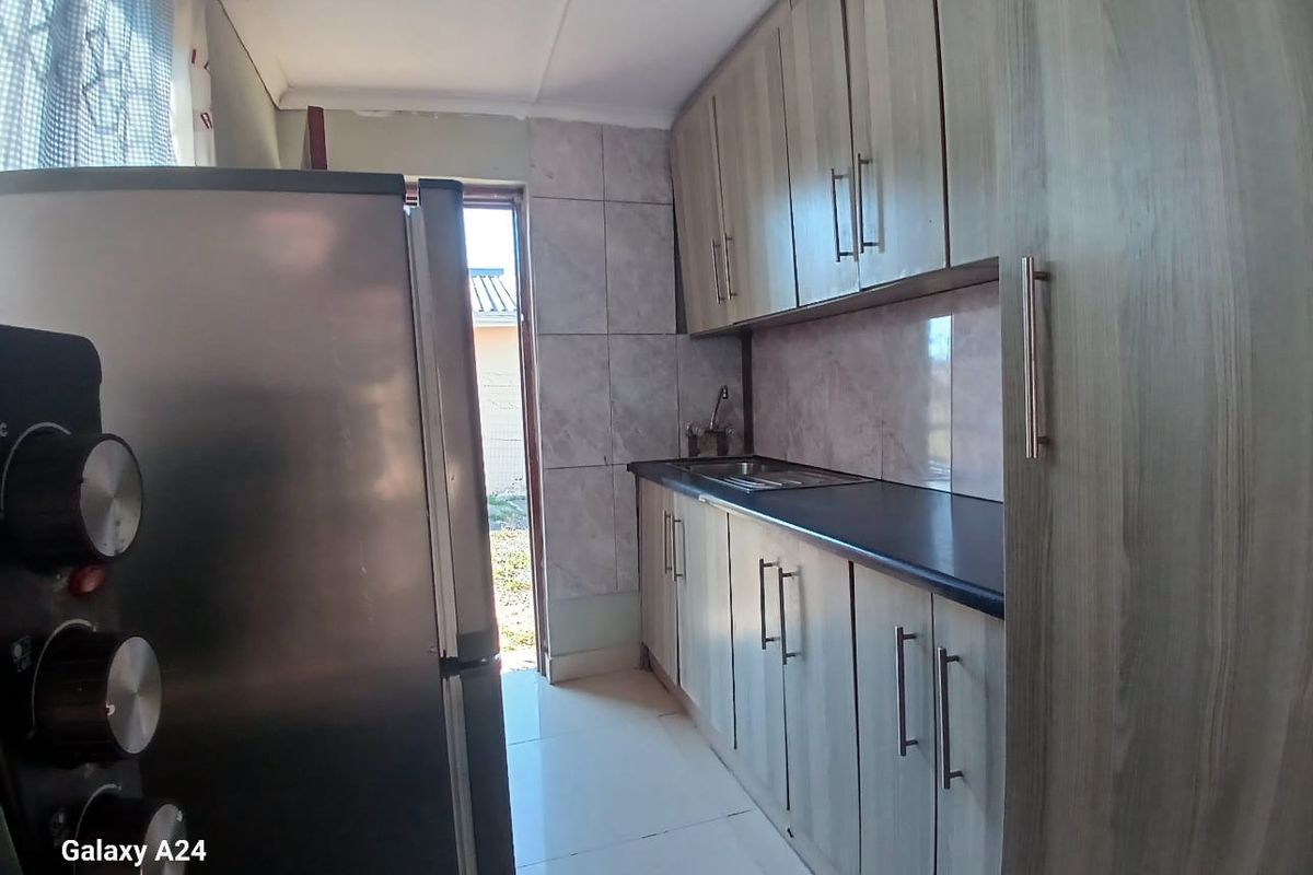 A compact kitchen featuring light wood-grain cabinetry, dark laminate countertops, tiled backsplash, and direct access to the exterior.