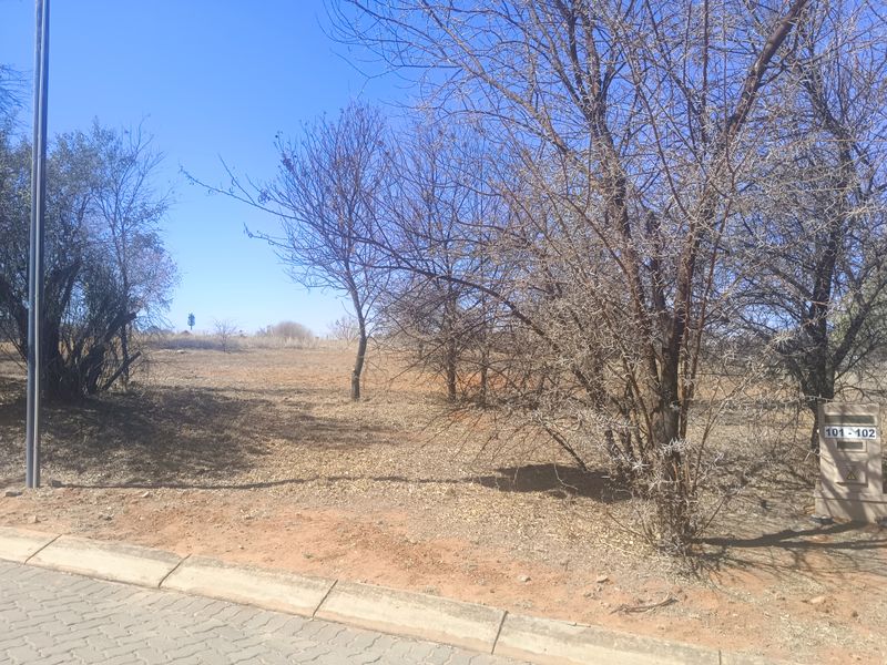 A vacant residential plot featuring natural vegetation, a paved curb, and a utility connection box at the street frontage.