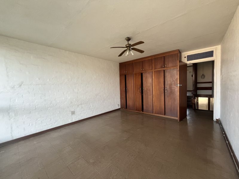 An empty bedroom featuring built-in wooden cupboards, a ceiling fan, and textured brick walls, offering a blank canvas for renovation.