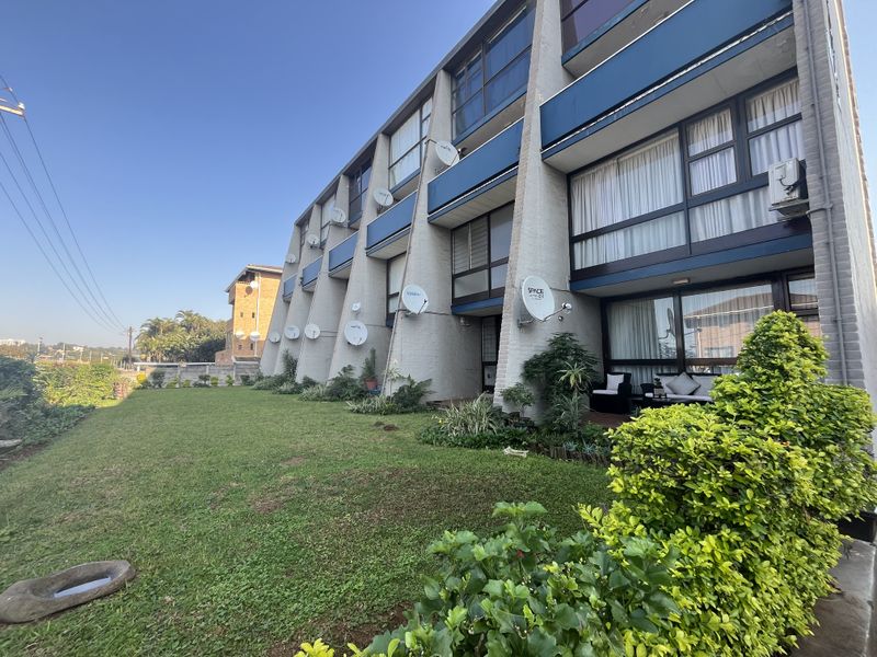 Rear view of a multi-story apartment building featuring a shared lawn, concrete structural pillars, and private ground-floor patio access with sliding glass doors.
