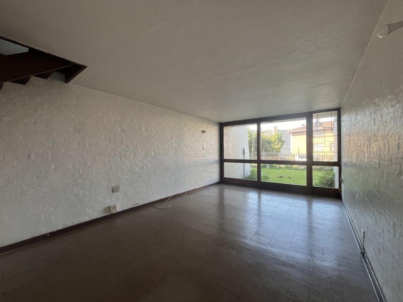 An empty living room featuring textured white brick walls, large windows overlooking the garden, and a staircase leading to the upper level.