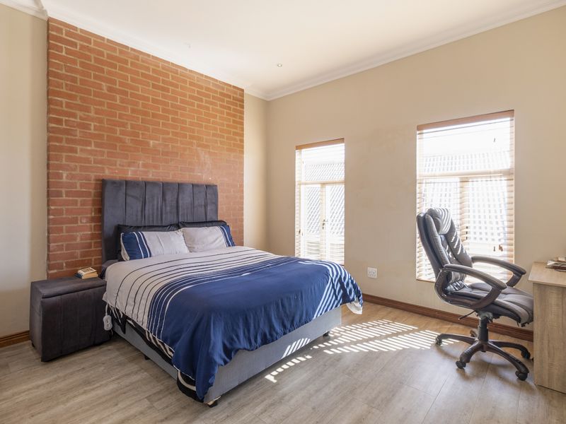 A well-lit bedroom featuring an exposed brick accent wall, light-toned laminate flooring, and dual windows with fitted venetian blinds.