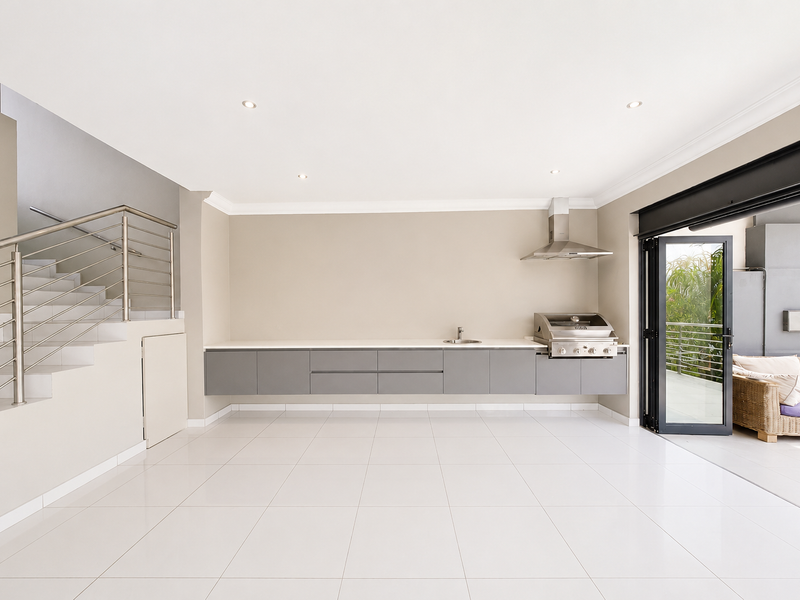 Spacious open-plan living area featuring a modern built-in braai, floating cabinetry, sleek tiled floors, and seamless access to the outdoor patio through bi-fold doors.