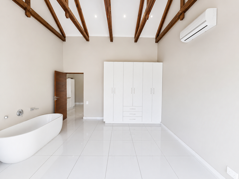 Spacious modern bathroom featuring a freestanding bathtub, built-in cabinetry, exposed timber ceiling beams, and sleek tiled flooring in a bright, neutral setting.