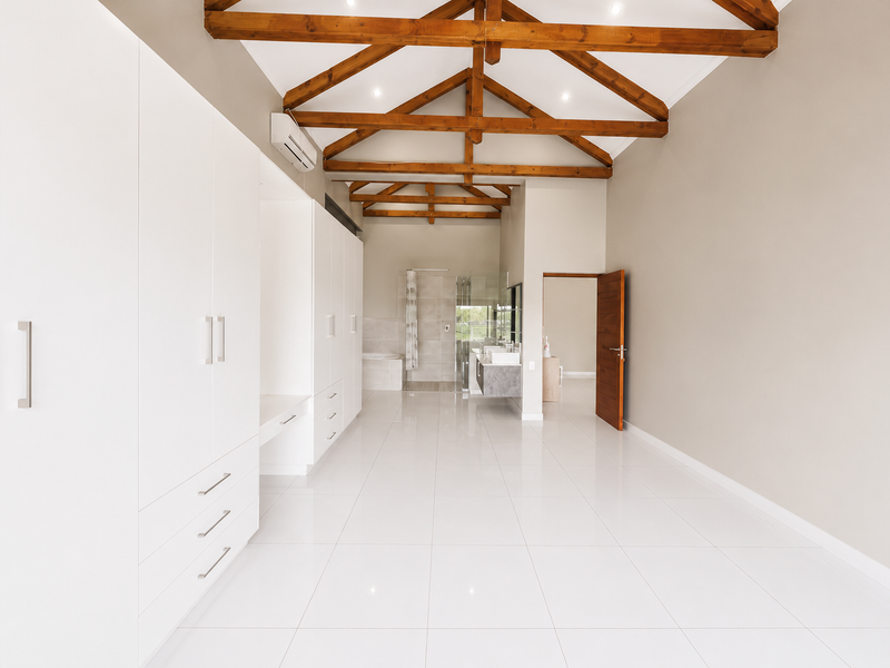 Spacious bedroom featuring high vaulted ceilings with exposed timber trusses, extensive built-in cabinetry, polished tile flooring, and direct access to an en-suite bathroom.