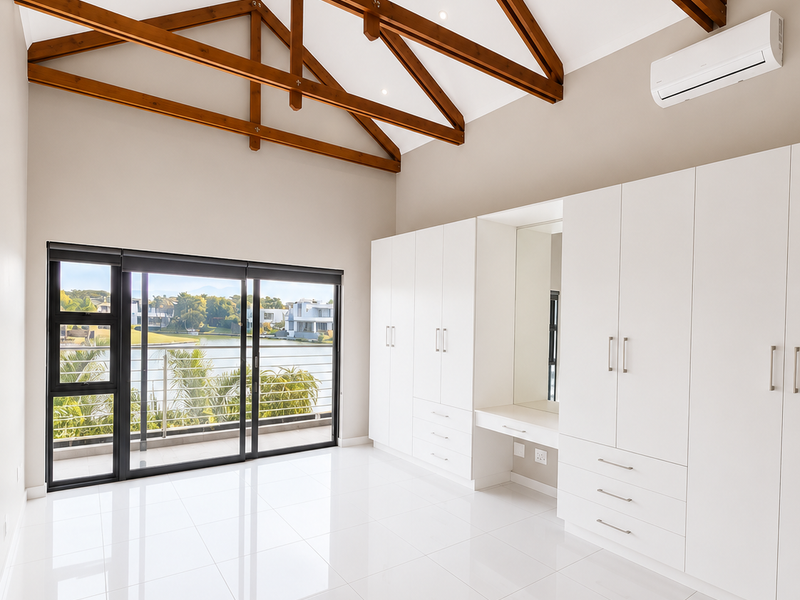 Spacious bedroom featuring high vaulted ceilings with exposed timber trusses, extensive built-in cabinetry with vanity, tiled flooring, and sliding doors opening to a balcony.