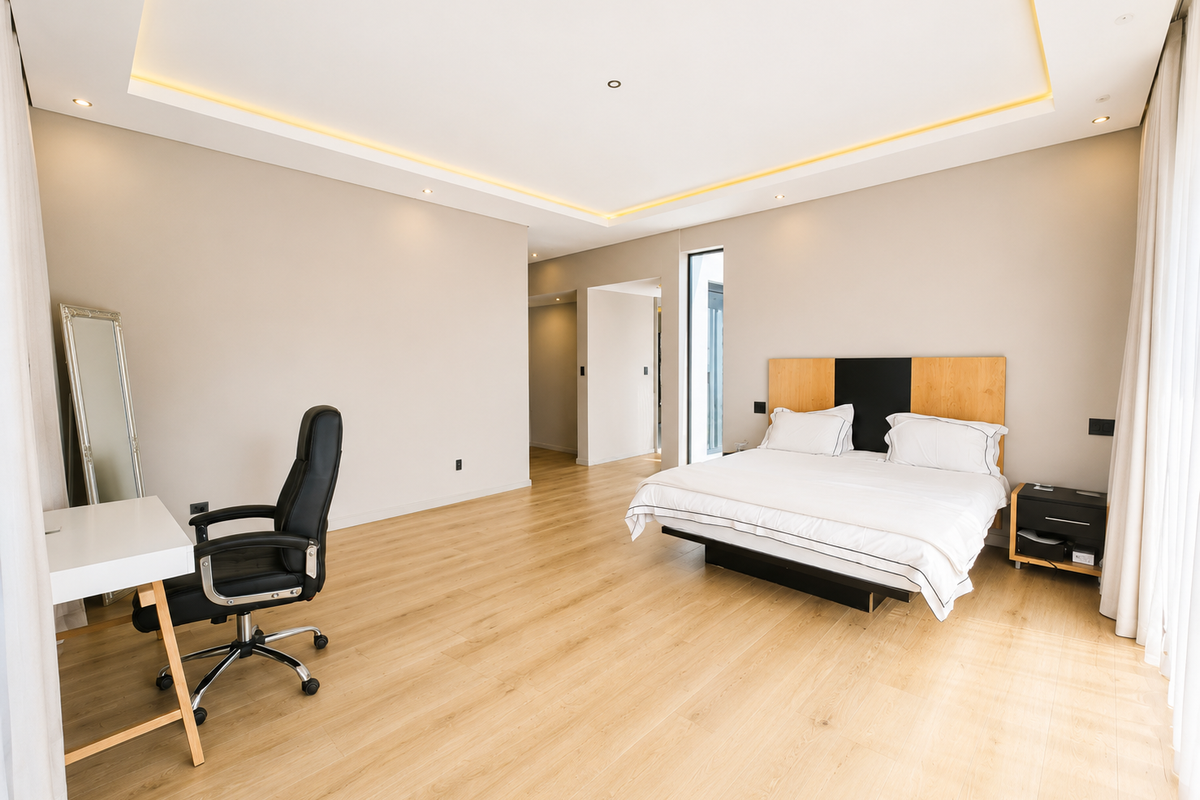Spacious modern bedroom featuring light wood-look laminate flooring, recessed ceiling lighting with ambient cove illumination, and a contemporary built-in headboard.