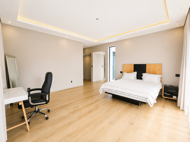 Spacious modern bedroom featuring light wood-look laminate flooring, recessed ceiling lighting with ambient cove illumination, and a contemporary built-in headboard.