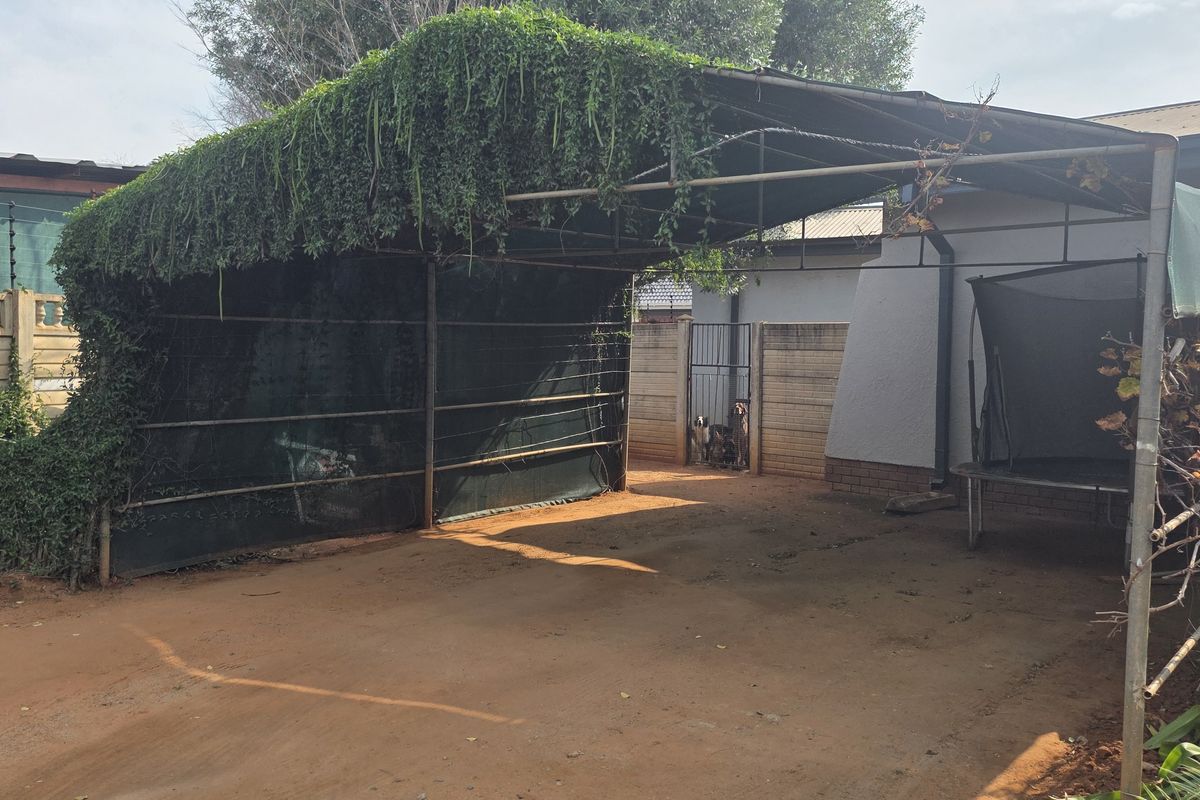 Rear exterior area featuring a metal carport structure with shade cloth, perimeter walling with electric fencing, and an unpaved driveway.