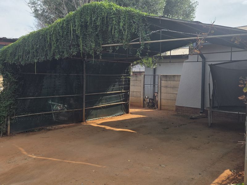 Rear exterior area featuring a metal carport structure with shade cloth, perimeter walling with electric fencing, and an unpaved driveway.