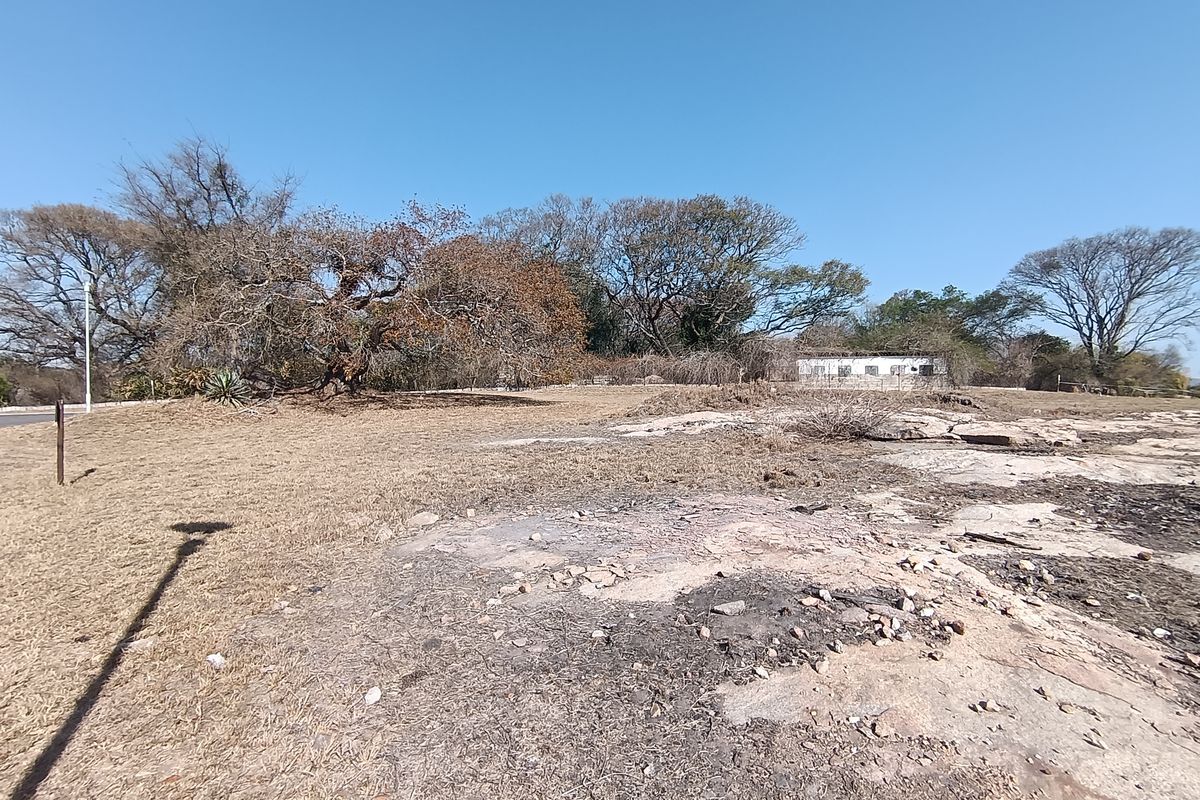 A view of a vacant residential plot featuring natural rocky terrain, mature trees, and perimeter fencing.
