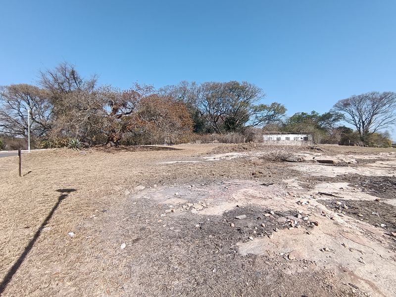 A view of a vacant residential plot featuring natural rocky terrain, mature trees, and perimeter fencing.