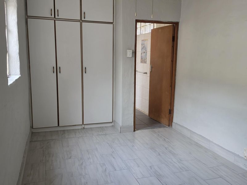 A clean, empty bedroom featuring built-in cupboards, a ceiling fan, and modern light-toned tiled flooring, with an adjacent doorway leading to a bathroom.