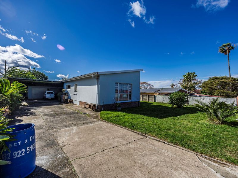 Rear view of the property featuring a concrete driveway, attached carport, and a spacious lawn area enclosed by brick boundary walls.