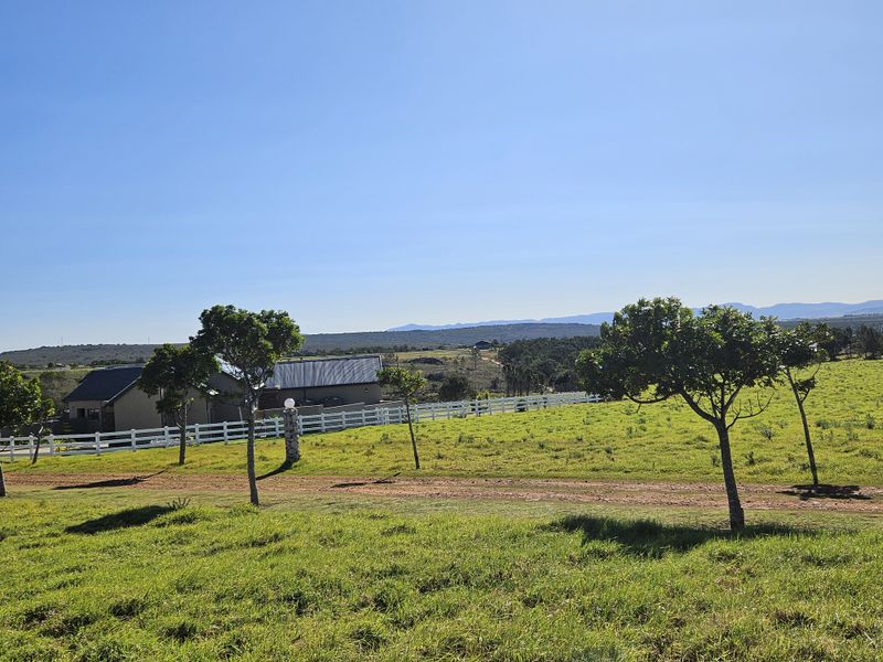 A scenic view of a vacant residential plot featuring a white picket fence, mature trees, and expansive rolling landscape under a clear blue sky.