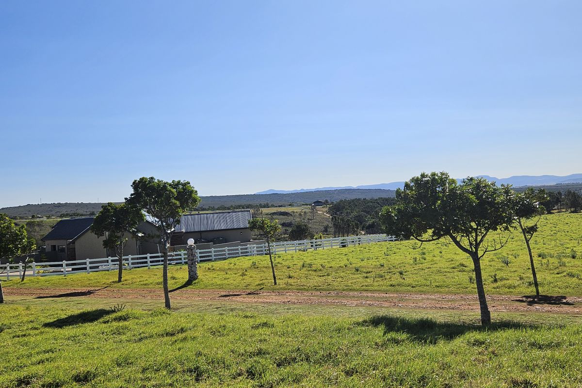 A scenic view of a vacant residential plot featuring a white picket fence, mature trees, and expansive rolling landscape under a clear blue sky.