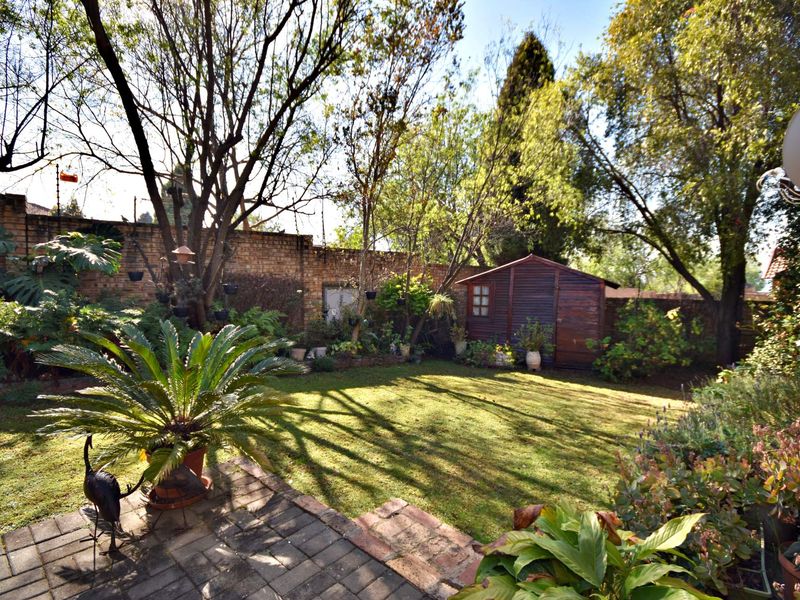 A private rear garden featuring a paved patio, established lawn, mature trees, and a wooden storage shed enclosed by a brick boundary wall.