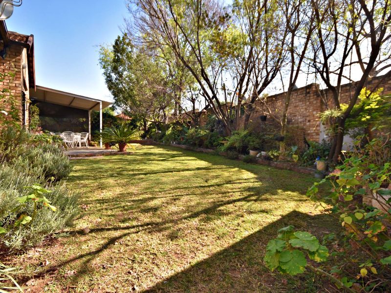 A private rear garden featuring a lawn, mature trees, a brick boundary wall, and a covered patio area.