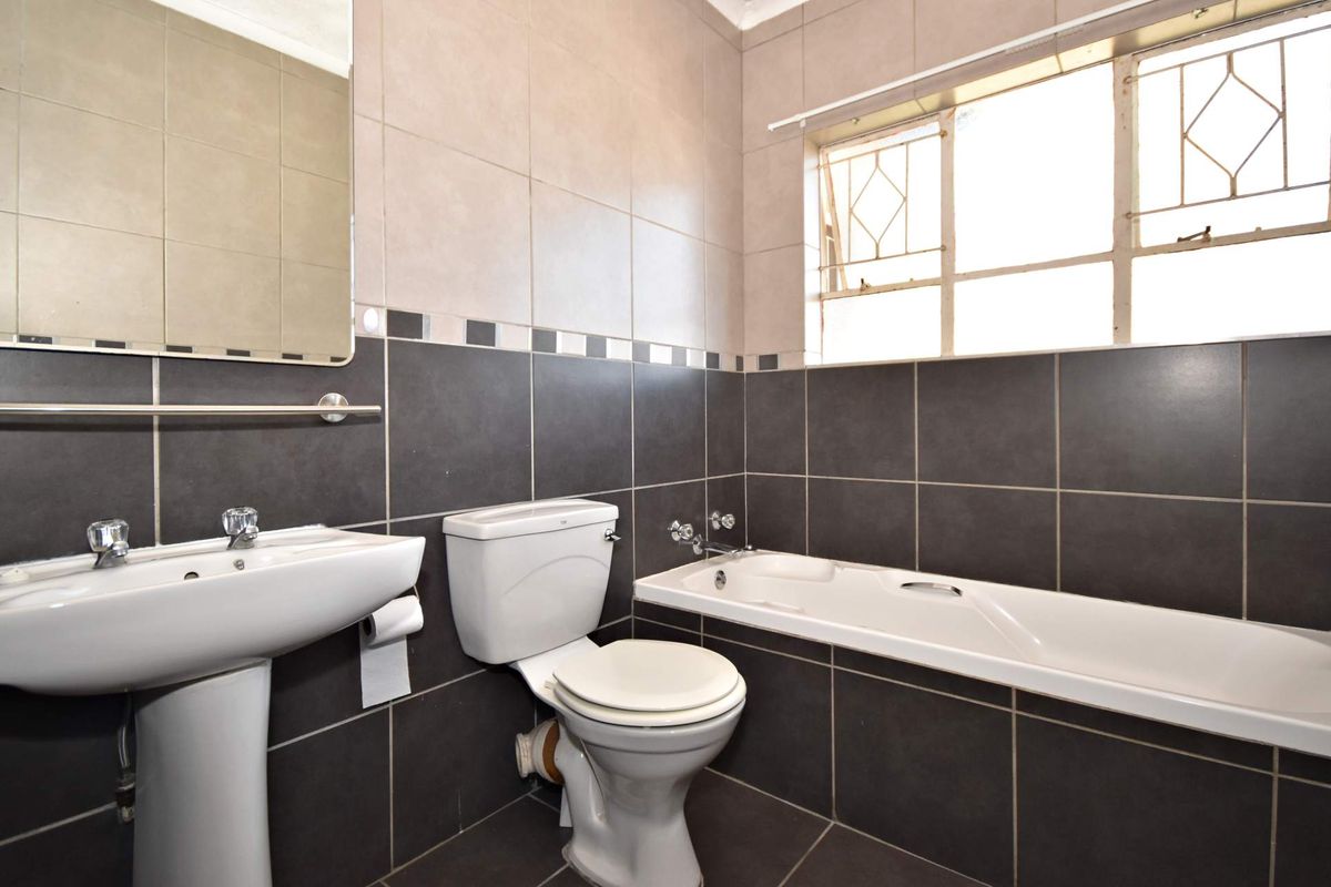 A functional bathroom featuring a pedestal basin, toilet, and bathtub with dark tiled walls and flooring, complemented by a window with security bars.