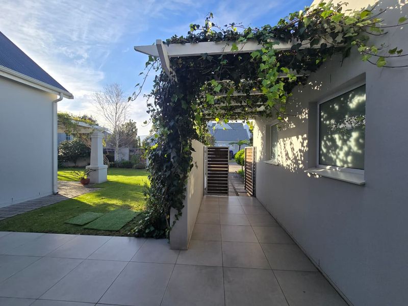 A covered patio area featuring a pergola with climbing greenery, tiled flooring, and a gated walkway leading to the garden.