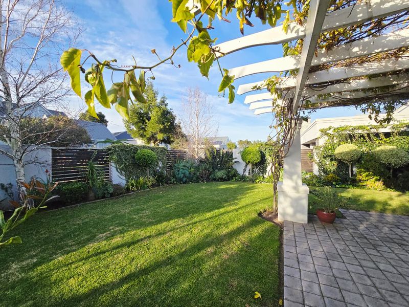 A well-maintained private garden featuring a paved patio area with a pergola, lush lawn, and established perimeter fencing.