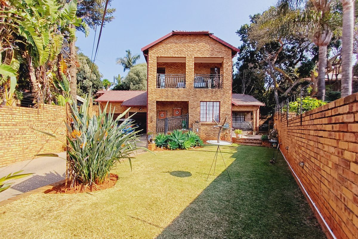 A well-maintained double-storey face brick house featuring an upper-level balcony, paved driveway, and a landscaped rear garden with established boundary walls.