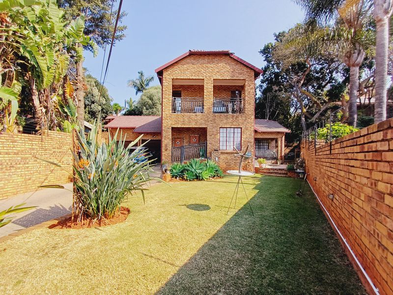 A well-maintained double-storey face brick house featuring an upper-level balcony, paved driveway, and a landscaped rear garden with established boundary walls.