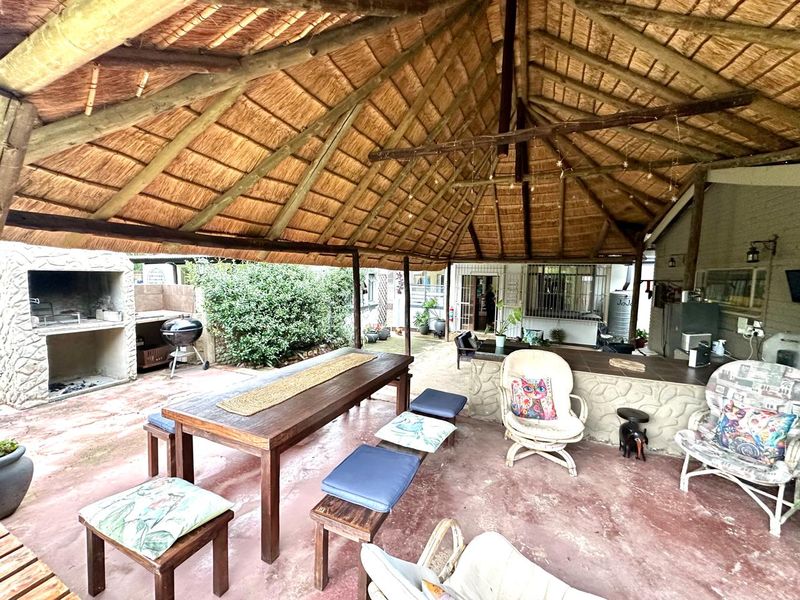 Covered outdoor patio featuring a traditional thatched roof, built-in braai area with stone cladding, and a spacious tiled counter, ideal for alfresco dining.