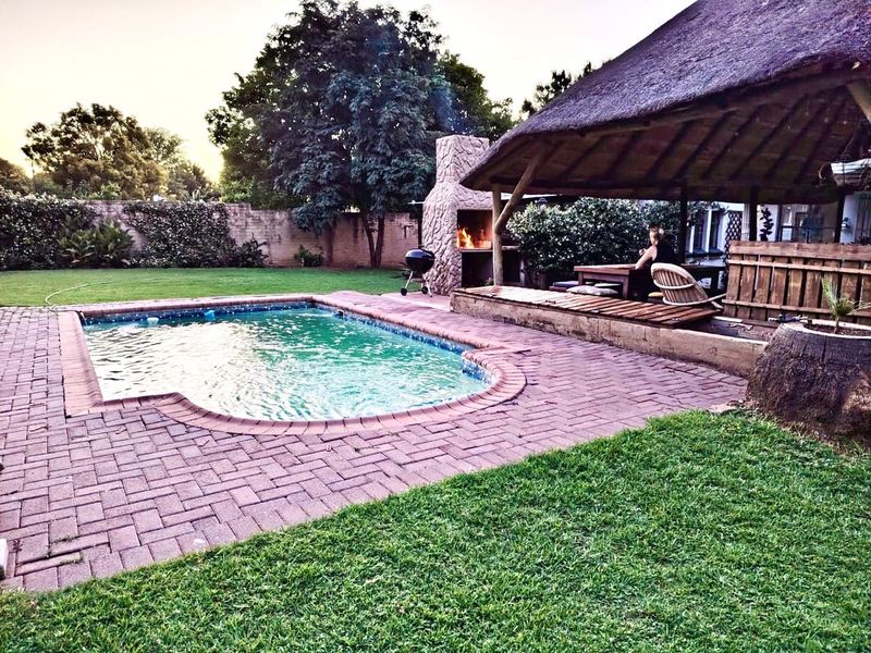 Outdoor entertainment area featuring a swimming pool, brick-paved patio, and a thatched lapa with a built-in braai, set within a private garden.