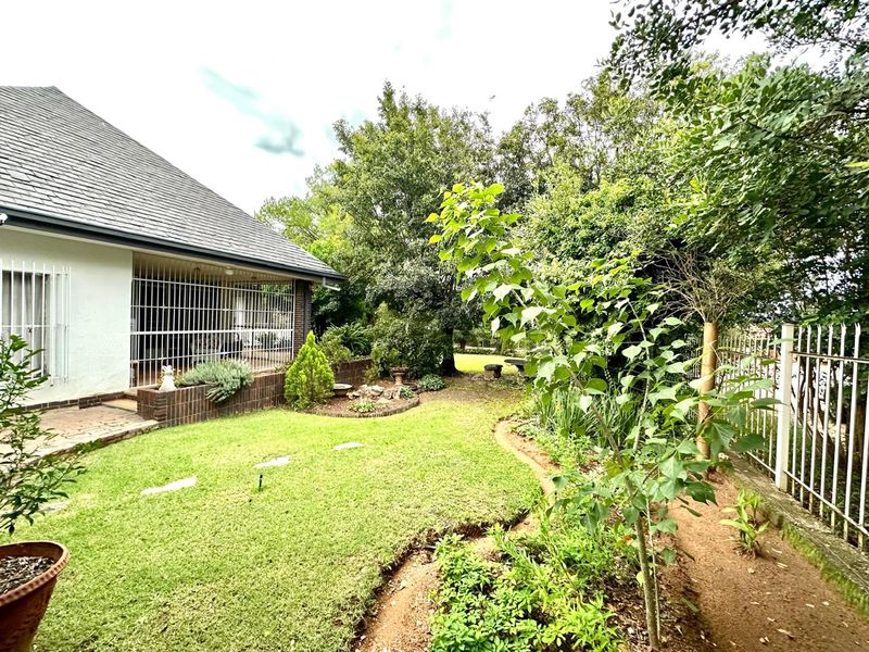 A view of the rear garden featuring a lawn, established trees, a brick planter, and a covered patio area with security bars.