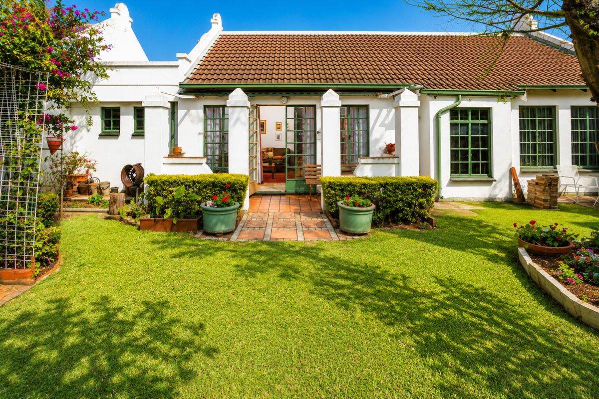 A well-maintained rear garden view featuring a white facade, terracotta roof tiles, green-framed windows, and a paved entrance leading into the home.