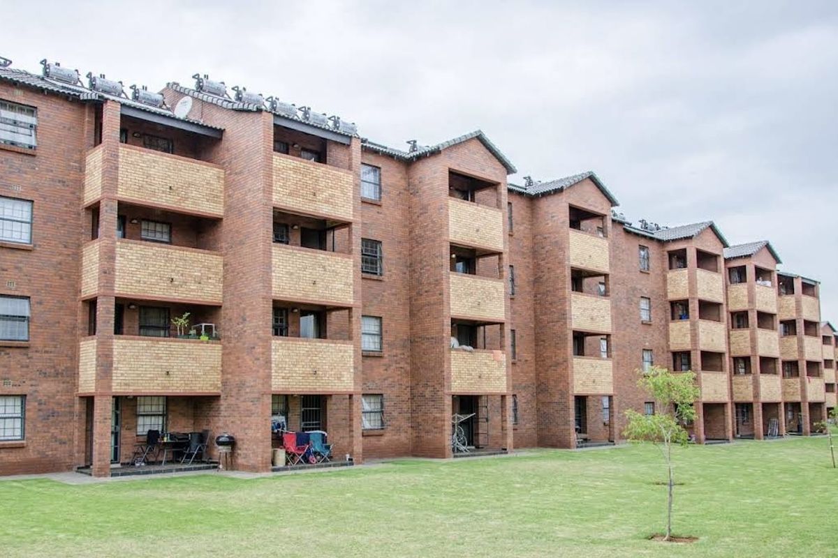 A multi-story apartment complex featuring face brick construction, private balconies for each unit, and solar water heating systems overlooking a communal garden area.