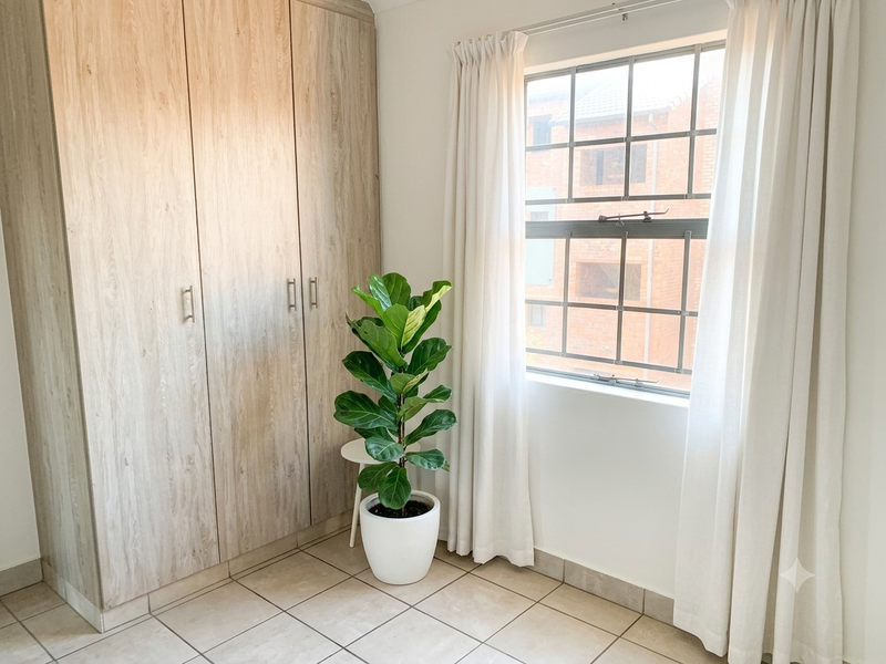 A bright, empty bedroom featuring light wood-finish built-in cupboards, neutral tiled flooring, and a window with security bars.