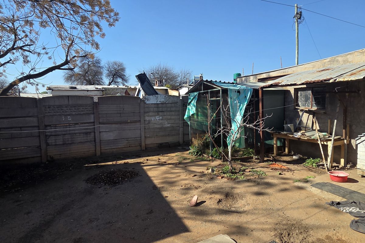 A rear yard area featuring a precast concrete boundary wall, a corrugated metal roof structure, and an unpaved ground surface.