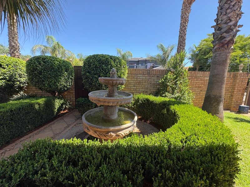 A well-maintained garden featuring a central stone fountain, manicured hedges, mature palm trees, and a brick boundary wall.