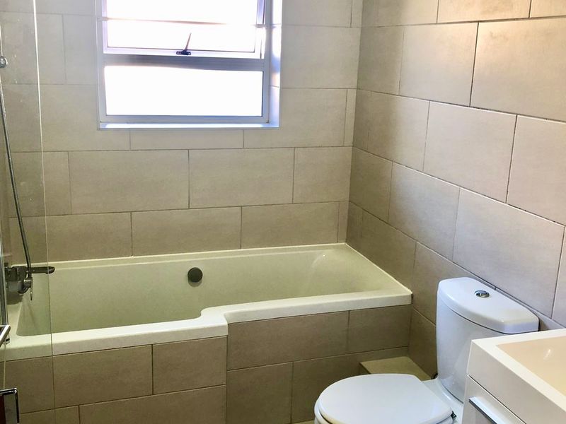 A clean, neutral-toned bathroom featuring a built-in bathtub with a glass shower screen, a toilet, and a vanity cabinet.