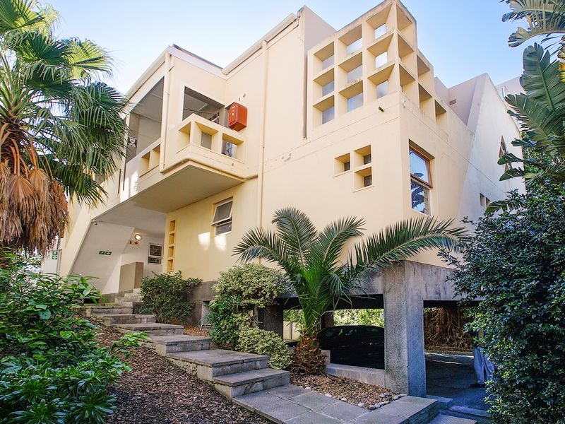 Exterior view of a multi-story apartment building featuring a concrete staircase, covered parking area, and architectural balcony details surrounded by established landscaping.