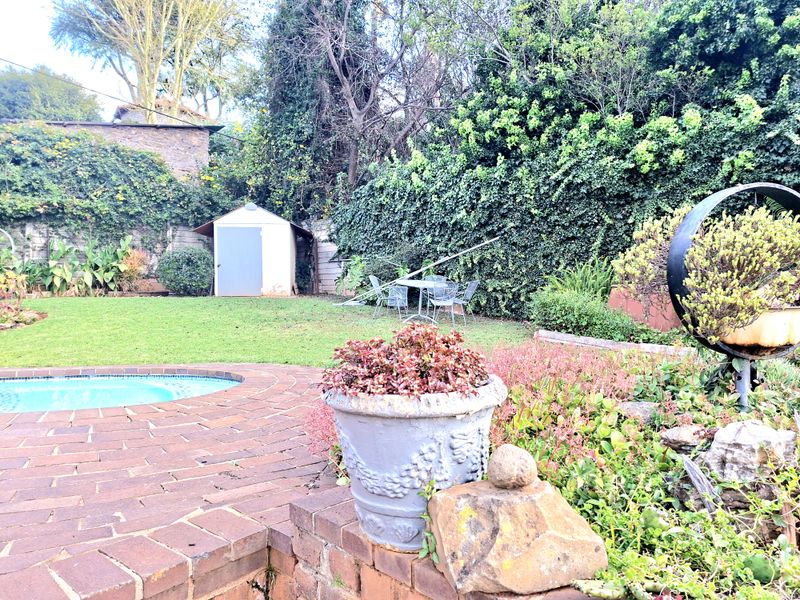 A private backyard featuring a swimming pool, brick paved patio area, established garden beds, and a detached storage shed.