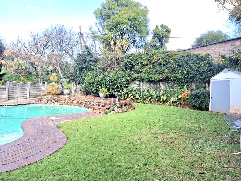 A private backyard featuring a swimming pool with brick paving, a stone retaining wall, landscaped garden beds, and a storage shed.